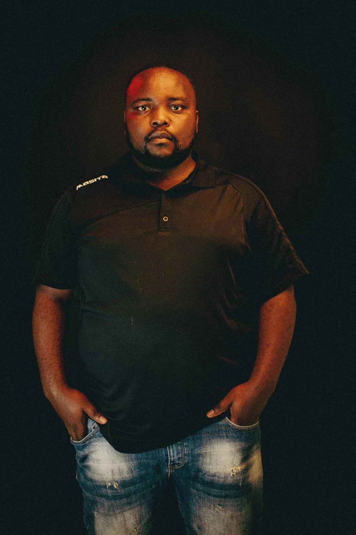 Photoreal digital twin of a man with short dark hair wearing a charcoal jacket over a black top and dark jeans against a deep green background.