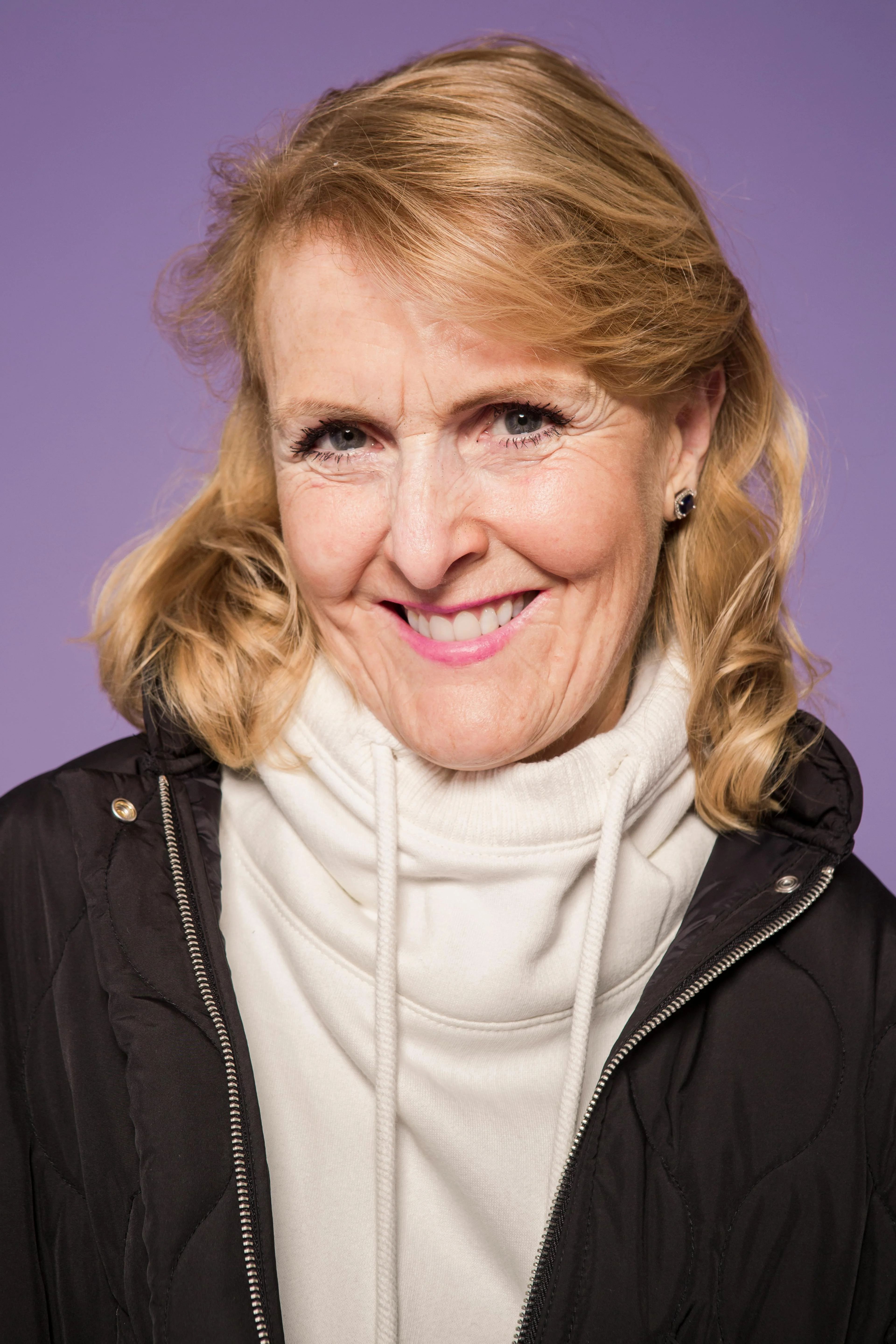 Photoreal digital twin of a woman with honey-blonde hair in a camel jacket over a peach top and ivory trousers against a mauve studio backdrop.